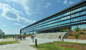 Library entrance shows metal and glass walls overlooking concrete steps and gathering students