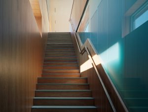 Stairway with exterior windows cast light down the wooden stairs and walls onto teal-tinted opaque glass