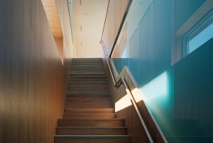 Stairway with exterior windows cast light down the wooden stairs and walls onto teal-tinted opaque glass