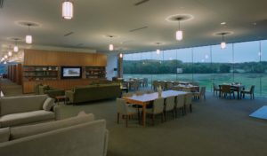 Blessings Golf Clubhouse dining room decorated in olive green chairs and couches and wooden tables with large windows that view the grass below at dusk