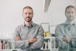 MBA team member headshot featuring Justin Hershberger with very short brown and beard wearing a gray shirt in front of an white office wall with arms crossed