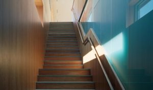 Stairway with exterior windows cast light down the wooden stairs and walls onto teal-tinted opaque glass