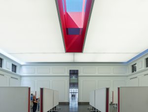 The red oculus skylight shines down to students working below in a large white room