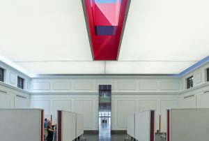 The red oculus skylight shines down to students working below in a large white room