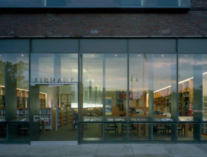 Gentry Public Library entrance of steel, brick, and glass looking in to modern wooden shelves filled with books