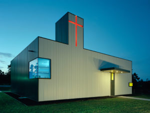 The church exterior seen at night, made of a box rib metal panel and red cross that shines from the top of a tower