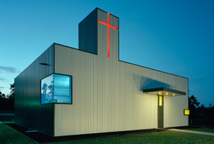 The church exterior seen at night, made of a box rib metal panel and red cross that shines from the top of a tower