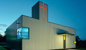 The church exterior seen at night, made of a box rib metal panel and red cross that shines from the top of a tower