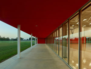 A sunset view from below the red stucco porch with green grass to the left and lit-up gym on the right
