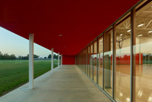 A sunset view from below the red stucco porch with green grass to the left and lit-up gym on the right