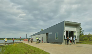 The stark metal Boathouse located along the water being used by park patrons as families on bike ride by