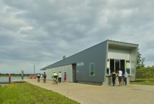 The stark metal Boathouse located along the water being used by park patrons as families on bike ride by