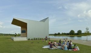 Families gather along the water next to a metal and wood angular building