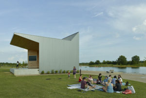 Families gather along the water next to a metal and wood angular building