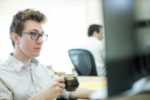Ethan Kaplan wearing a patterned shirt and glasses drinking a coffee and reading his monitor