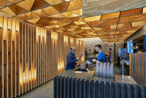 Geometric wooden ceilings and vertical wood slatted walls surround a modern bar with a patron ordering a drink