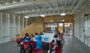 Children gather in a stark white room with wooden modern stairs and polished concrete floors