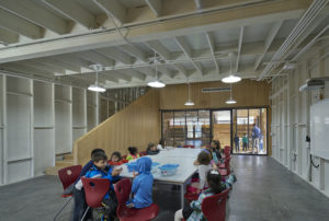 Children gather in a stark white room with wooden modern stairs and polished concrete floors