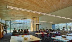 The Kids Circle gathers in a Lamplighter classroom with glass windows to the exterior, wooden walls and ceilings, and lights modern lights hanging from the ceiling