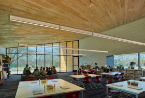 The Kids Circle gathers in a Lamplighter classroom with glass windows to the exterior, wooden walls and ceilings, and lights modern lights hanging from the ceiling