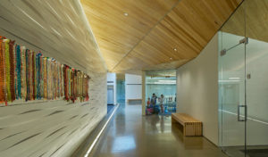Hallway showing geometric angled wooden roof, white walls, glass doors and windows, and woven art handing on the walls