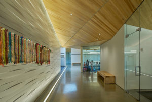 Hallway showing geometric angled wooden roof, white walls, glass doors and windows, and woven art handing on the walls