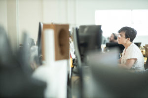 A young female architect with short dark hair, glasses, and a white shirt working diligently at a desk with two monitors in a white office space with bright natural light