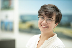 MBA team member headshot featuring a young female architect with short dark hair, glasses, and a white shirt smiling in front of a white office space with architectural images mounted on the walls