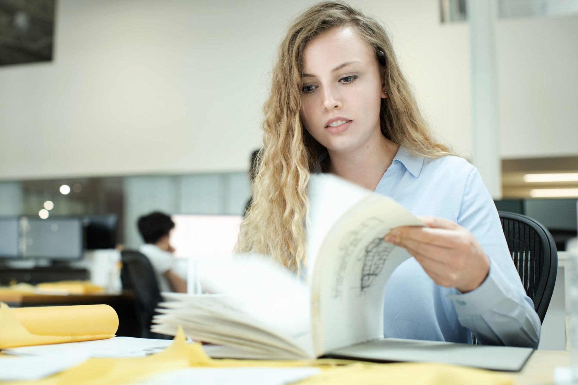 A young female architect with curly blonde hair wearing a bright blue shirt working at her desk flipping through architecture plans in a white office