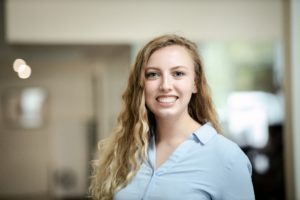 MBA team member headshot featuring a young female architect with curly blonde hair smiling in front of a white office wall