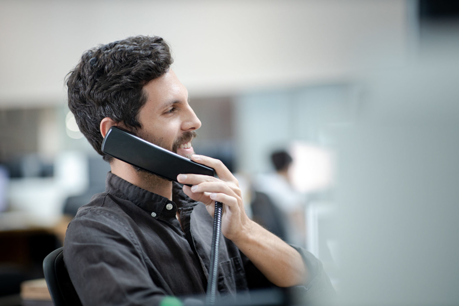 A male architect with short dark hair and beard wearing a dark gray shirt sitting in an office chair on the phone laughing in a bright white office space