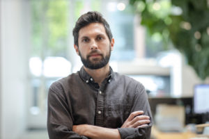 MBA team member headshot featuring a male architect with short dark hair and beard wearing a dark gray shirt standing with arms crossed in front of an out of focus white office with green plants