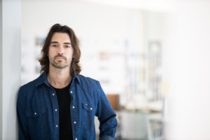 MBA team member headshot featuring a male architect with long brown hair and a trimmed beard wearing a black shirt under an unbuttoned dark blue denim shirt standing in front of a white office space