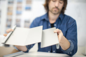 A male architect with long brown hair and a trimmed beard wearing a black shirt under an unbuttoned dark blue denim shirt working at a desk folding a 3d model and examining his work