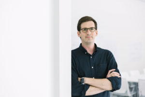 MBA team member headshot featuring A male architect with dark hair wearing round glasses and a deep navy button-up shirt and rolled up sleeves with crossed arms leaning on a wall in a white office