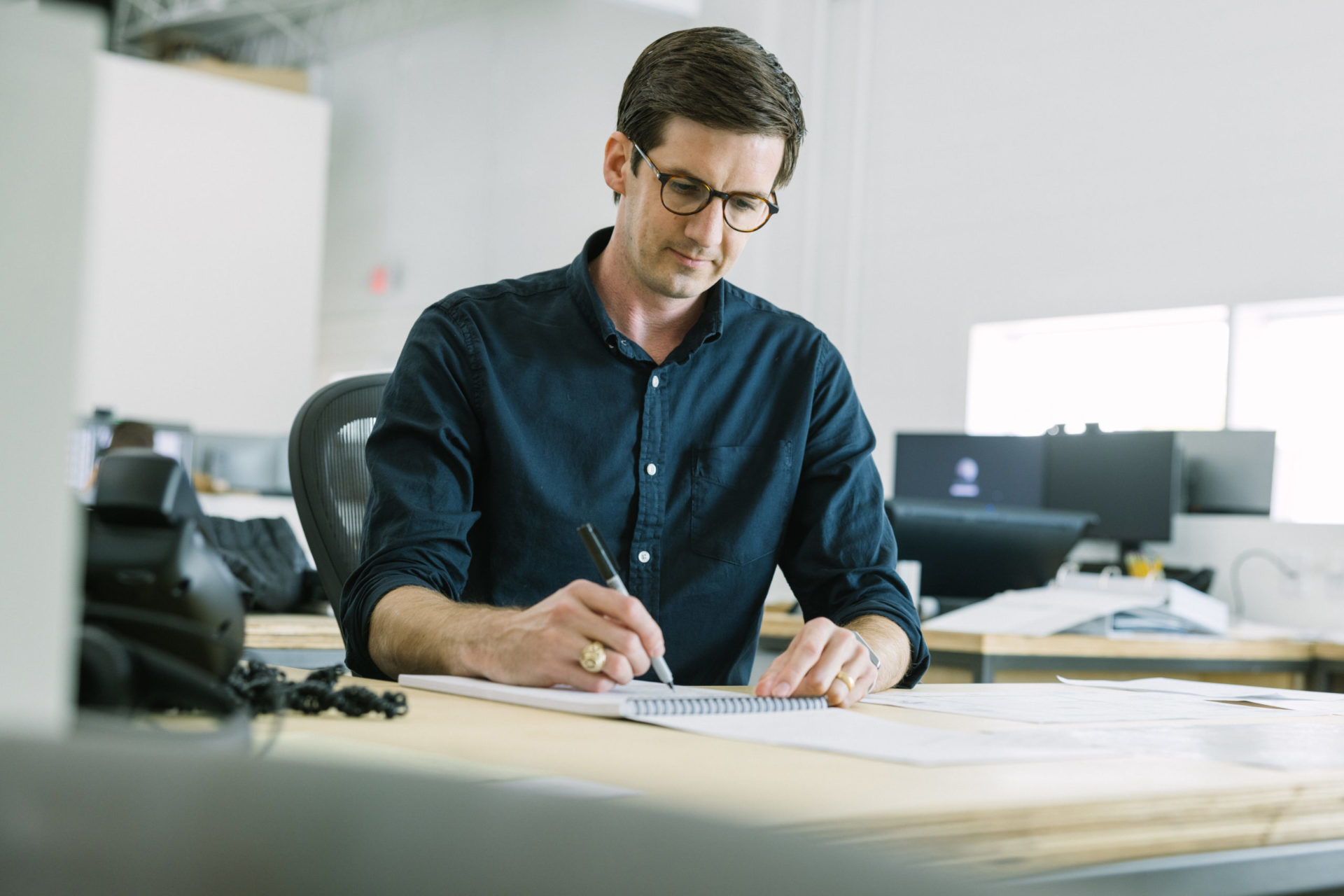 A male architect with dark hair wearing round glasses and a deep navy button-up shirt and rolled up sleeves working at a desk in a modern office