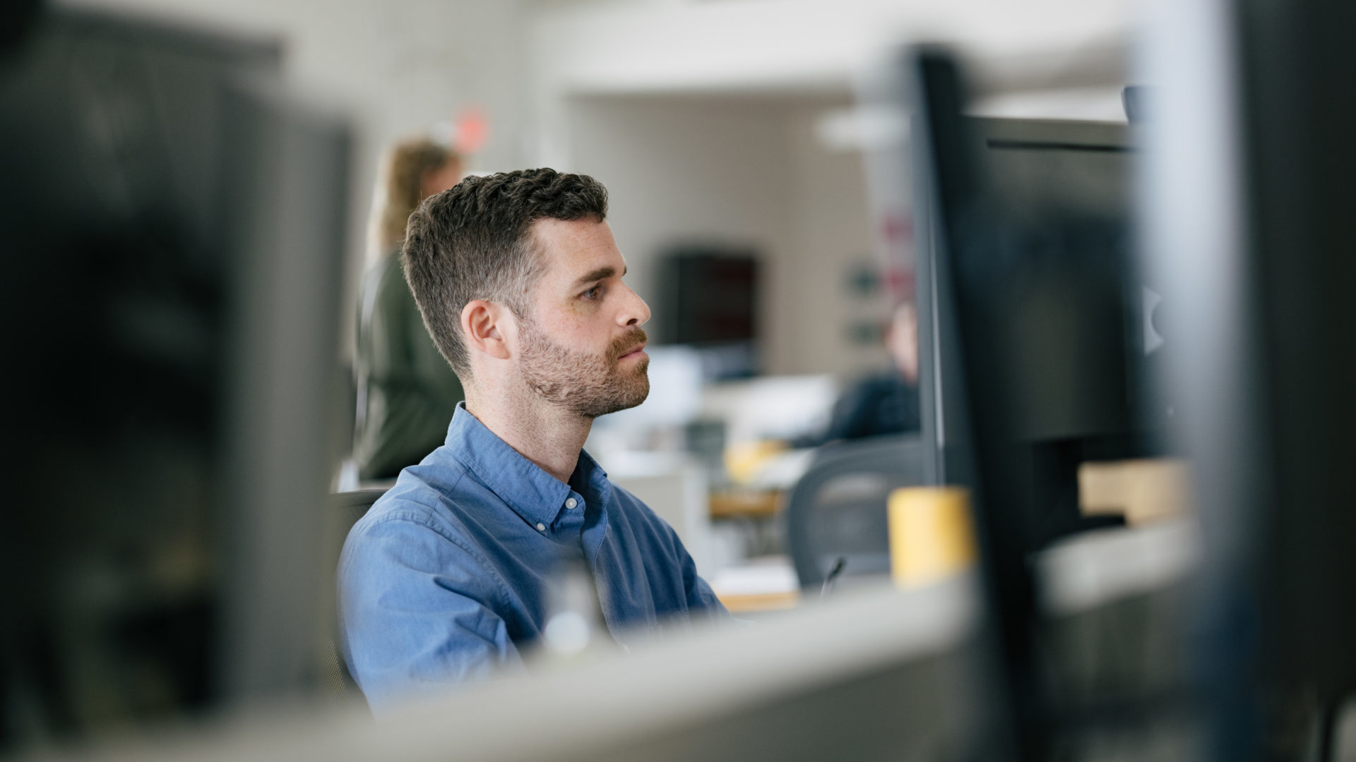 A male architect with short gray and brown hair and neatly trimmed beard wearing a light blue shirt working at his desk with eyes focused on his computer monitor