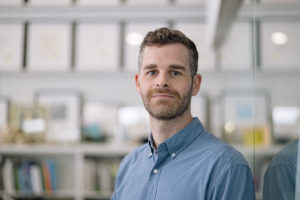 MBA team member headshot featuring a male architect with short gray and brown hair and neatly trimmed beard wearing a light blue shirt leaning on a glass wall in front of a white office space