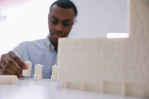 An out of focus young male architect wearing a light blue shirt working carefully with scale architectural models in a white office