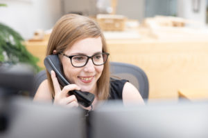 A young woman with red hair and glasses wearing a black tank top answering the phone at a desk