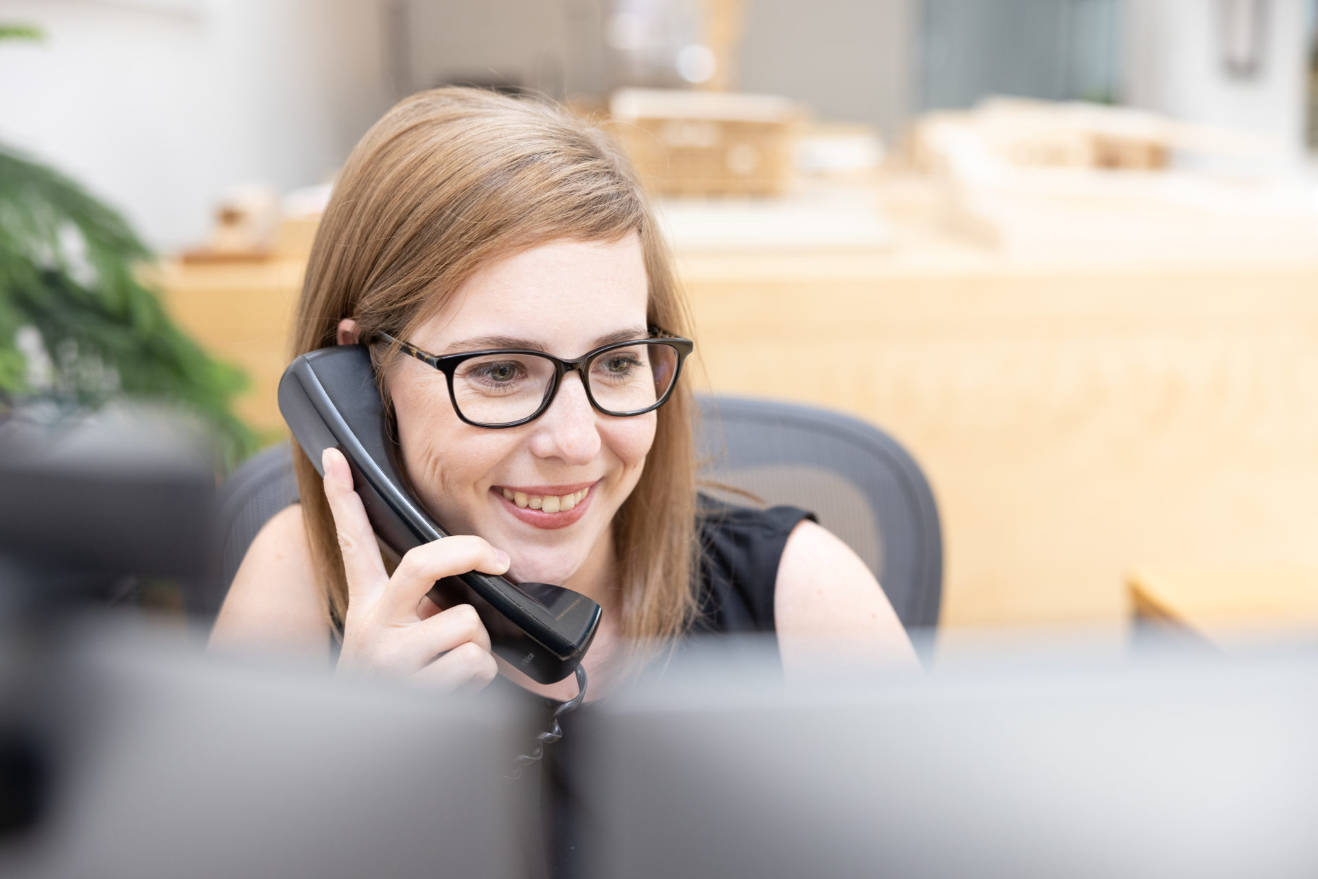 A young woman with red hair and glasses wearing a black tank top answering the phone at a desk