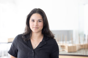 MBA team member headshot featuring a young female architect with dark hair wearing a black shirt standing in front of architectural scale models and white office wall