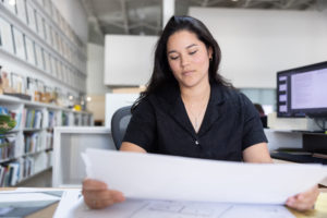 A young female architect with dark hair wearing a black shirt working at a desk reviewing architectural plans
