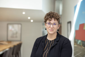 MBA team member headshot featuring a young female architect with curly brown hair and round glasses standing in a white office with large architectural images on the walls behind her