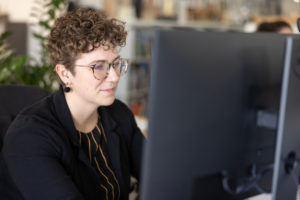 A young female architect with curly brown hair and round glasses working on a computer at a desk in the MBA office